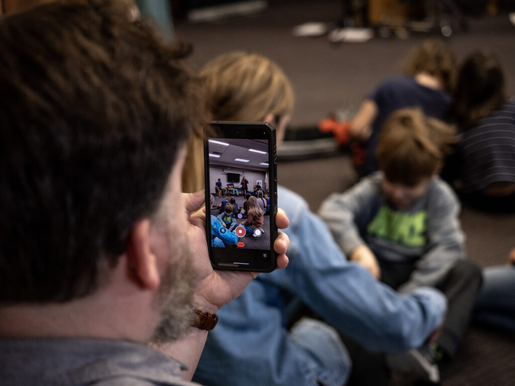 A man tapes a performance of a singer on his phone. The setting is a bright room with an audience of children blurred in the background.