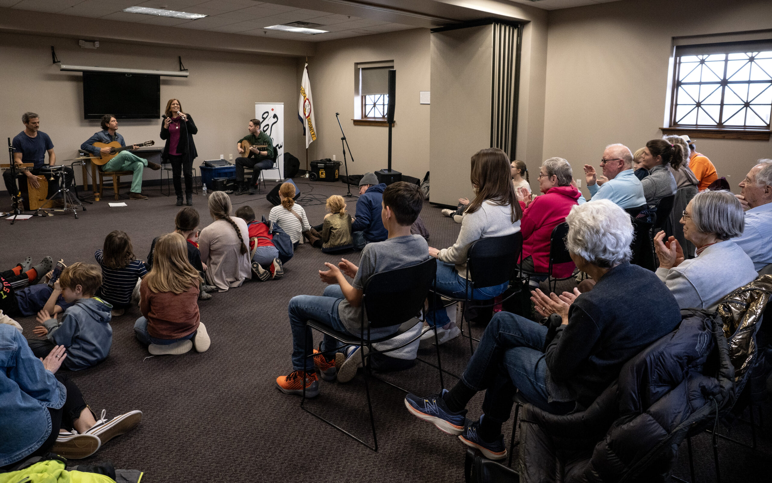 A woman performs with her band to an audience of onlookers of all ages at a public library's presentation room