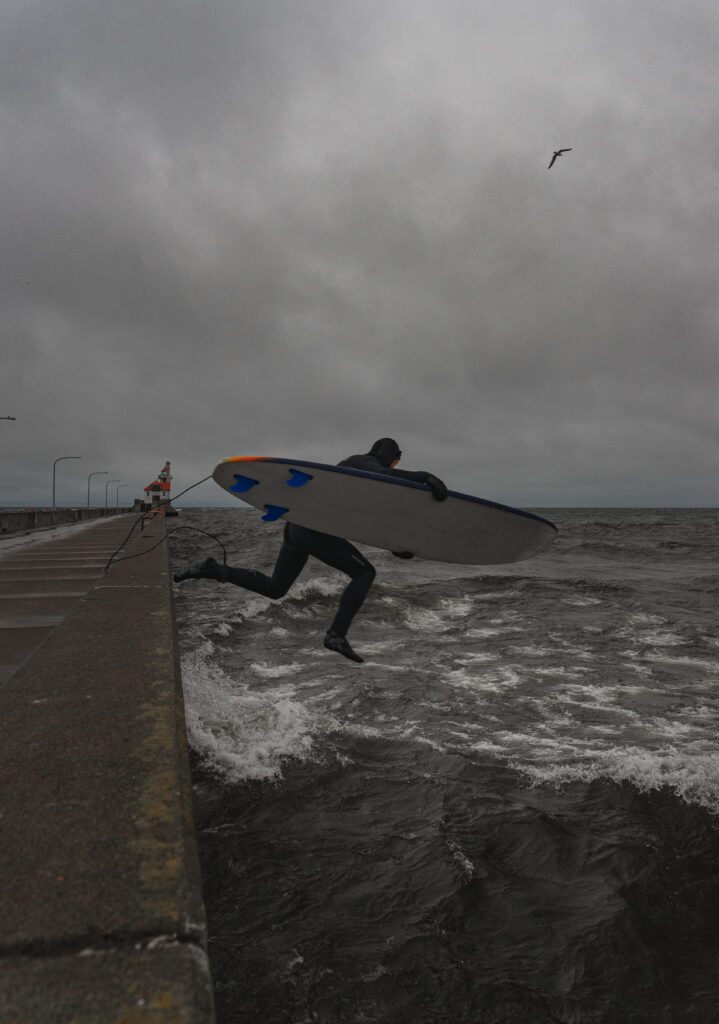 A surfer in a wet suit, holding their surf board jumps into a lake from the ledge of a concrete harbor.