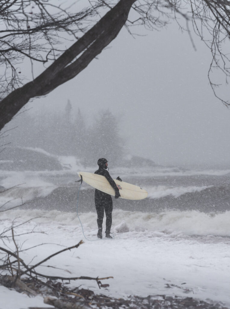 A surfer wearing a wet suit and holding a pale yellow board looks out at the waves in the lake from a snowy shore.