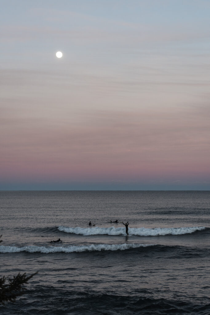 A view of the lake and horizon with pink skies. There are several surfers in the water.
