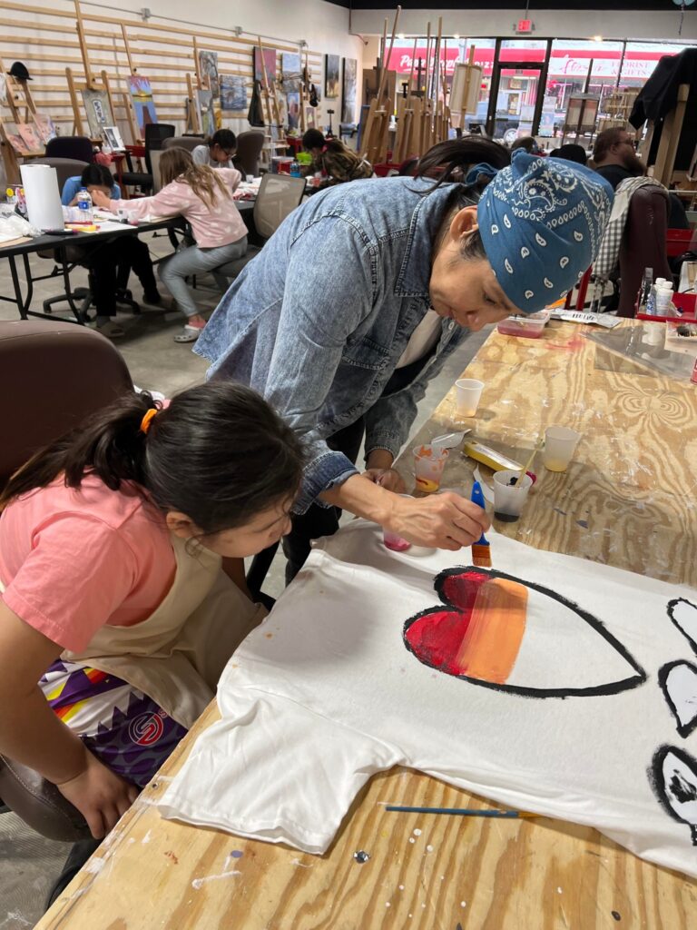 A woman with medium light skin paints a heart on a t-shirt as a young girl watches