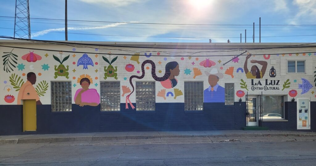 A mural on the side of a building that reads La Luz Central Cultural. Faceless people of medium and dark skin tones and colorful objects are represented.
