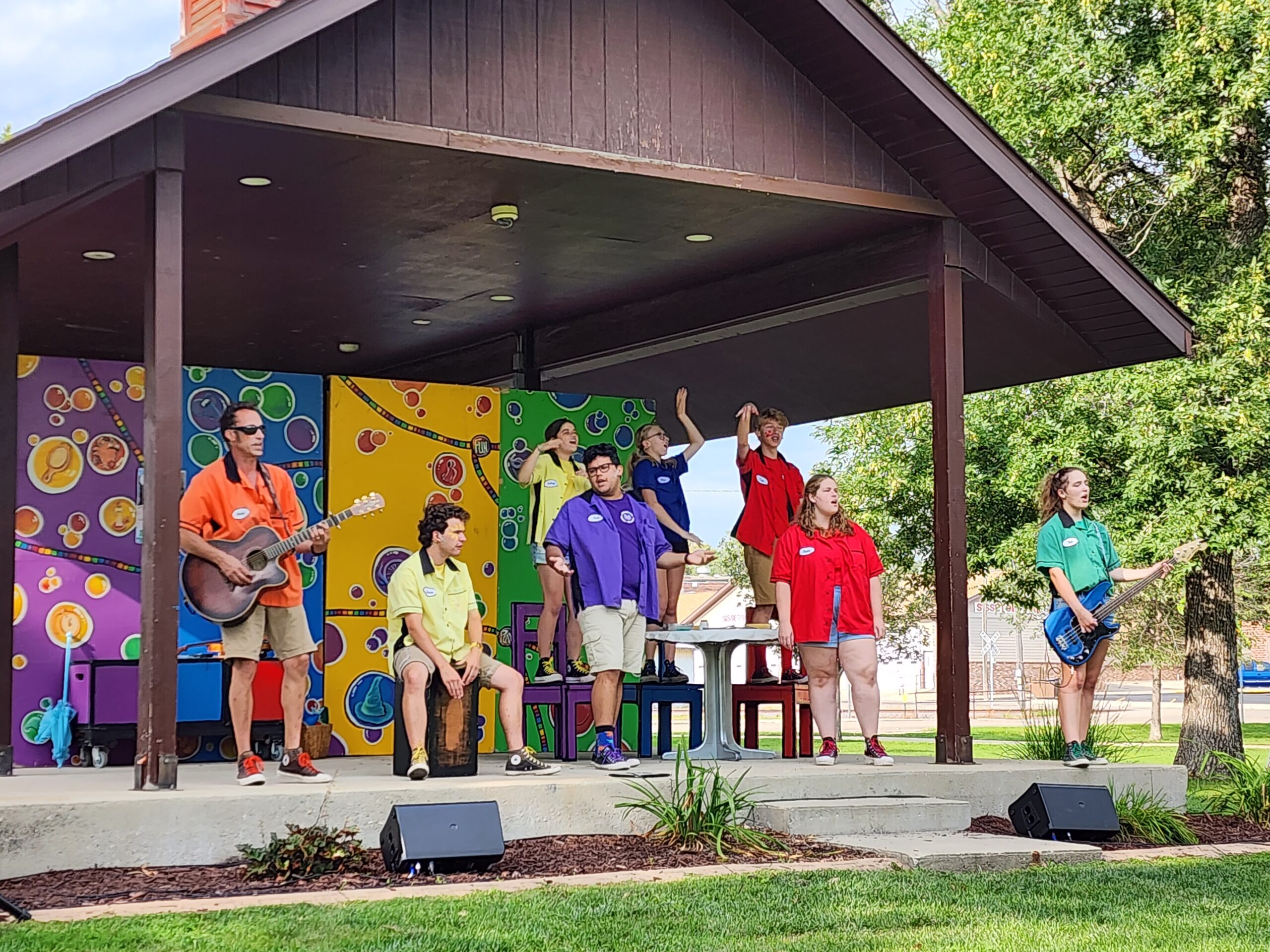 People in colorful shirts perform outside in front of a colorful mural.