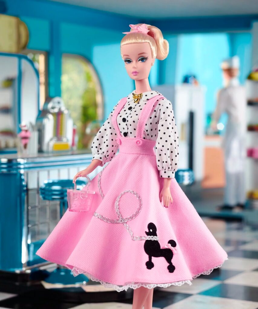 A Barbie doll with a polka-dotted blouse and large pink poodle skirt, standing in front of a soda shop backdrop.