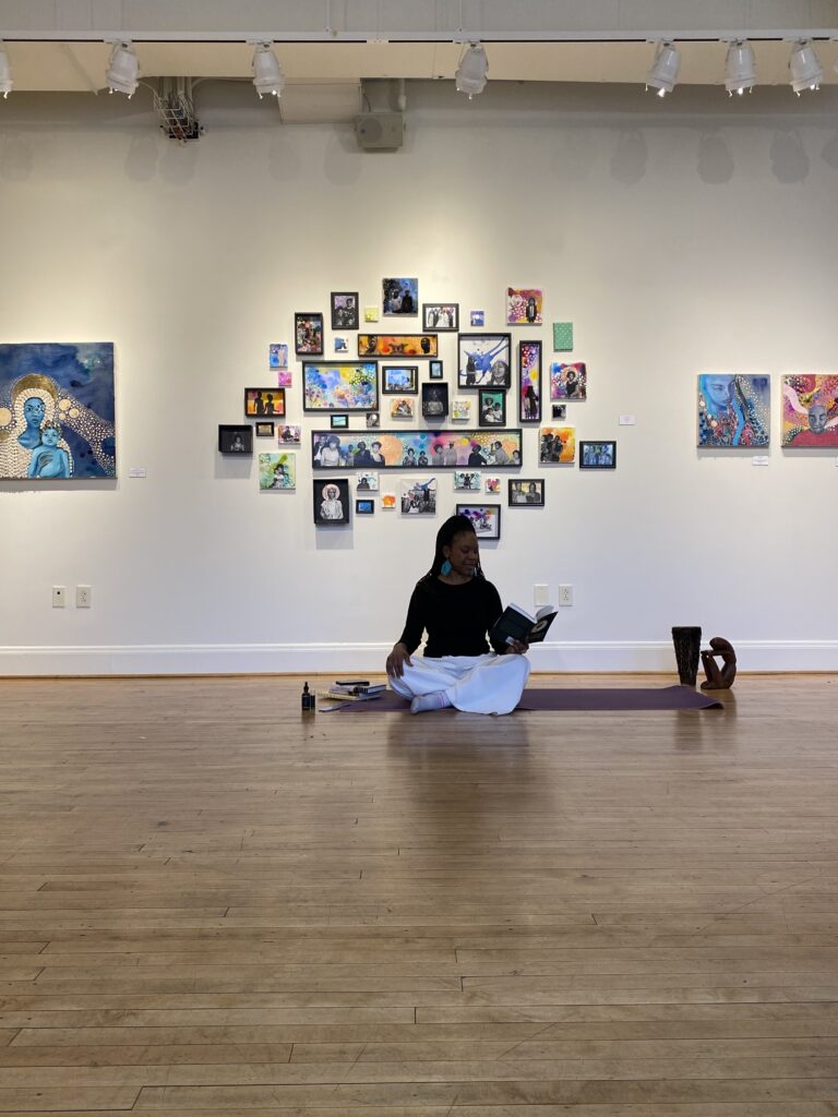 A person of dark skin tone sitting on the floor in an art gallery and holding a book, with a collection of artwork behind them.
