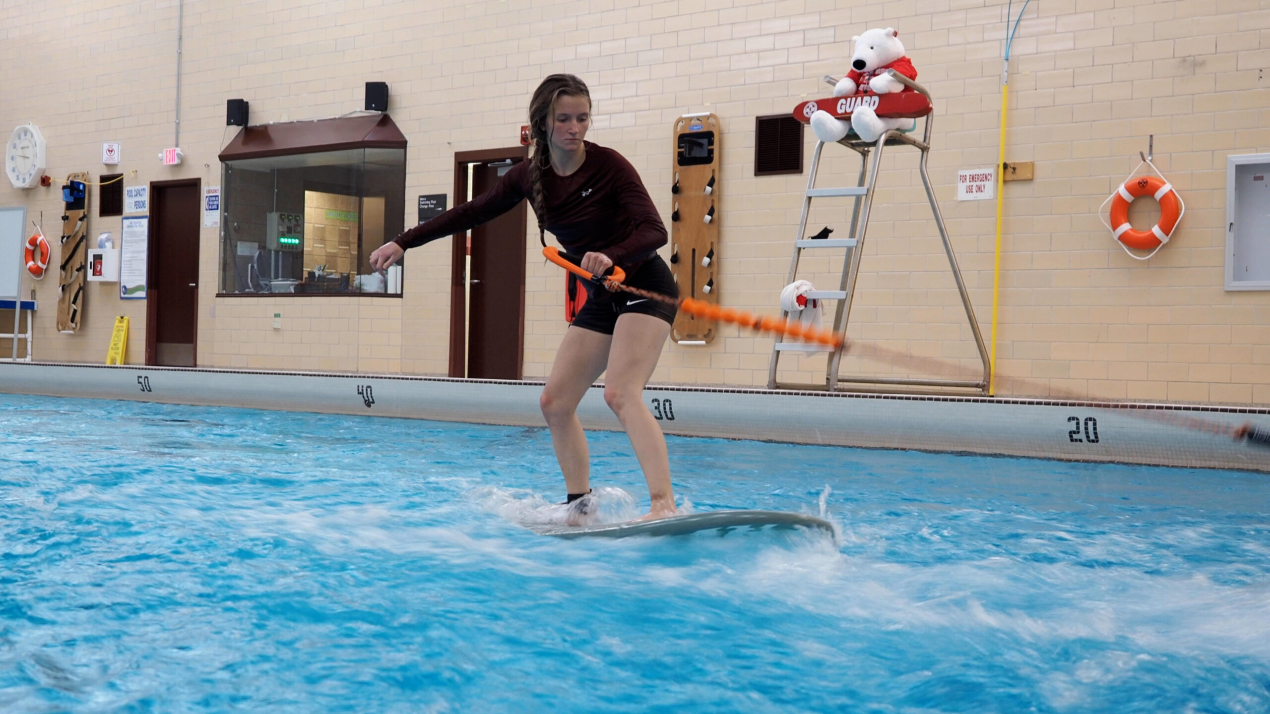 A person stands on a surf board and balances on simulated waves in a swimming pool. They are holding a cable with one hand as they balance their body.