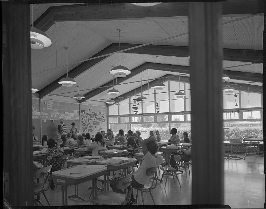 Students sitting at their desks in a classroom. It has tall ceilings with large beams and large windows.