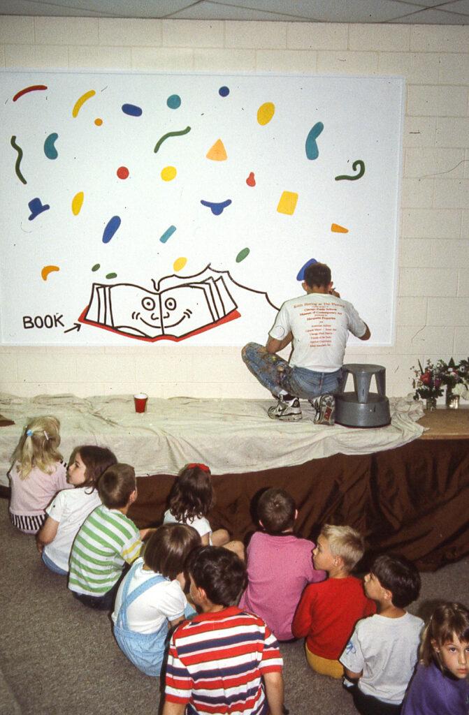 About ten children sit in two rows in front of an elevated platform where someone is crouching with their back to the children to paint a large thought bubble over an open book that is smiling.