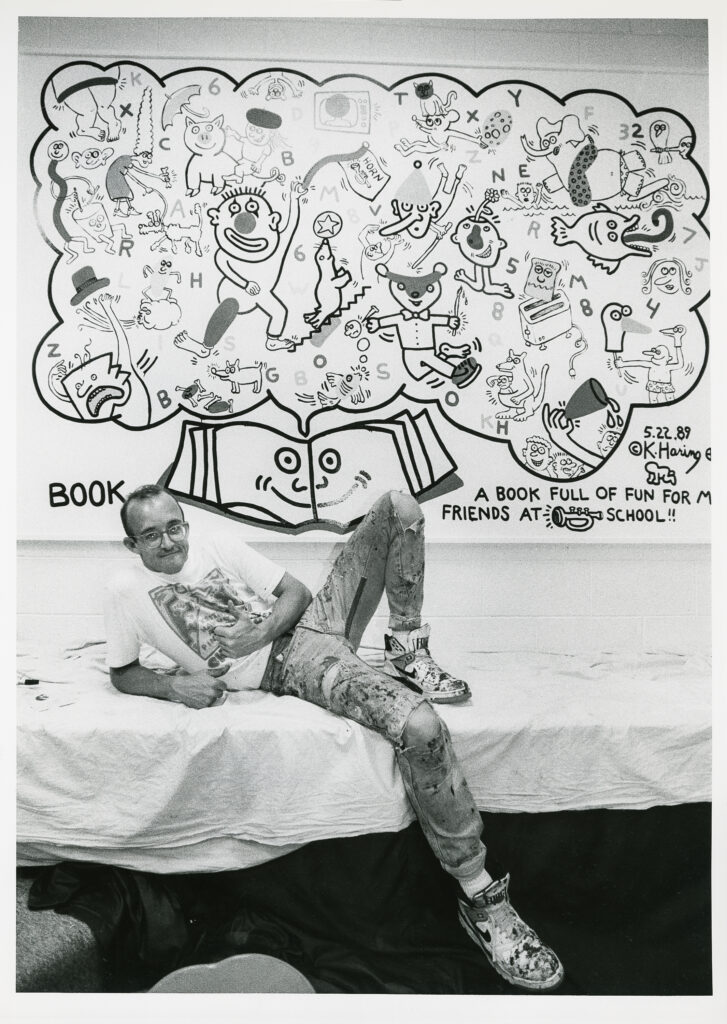 A black and white image of someone lying propped on their elbow on a table covered in a white sheet. Behind them is a mural with a smiling open book below a thought bubble containing playful imagery of people and animals.