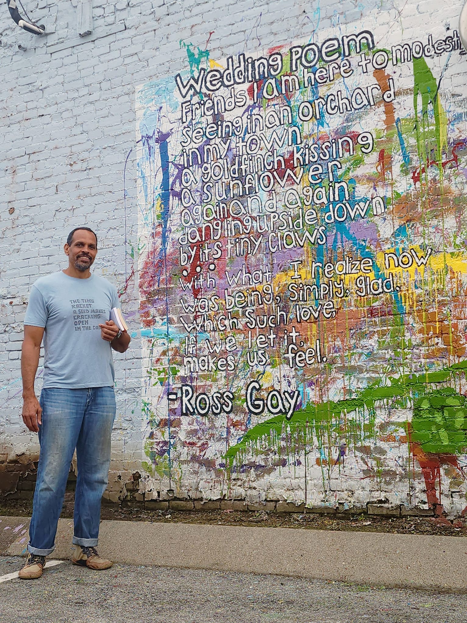 A man of medium light skin tone poses in front of a colorful mural and words from his poem Wedding Poem.