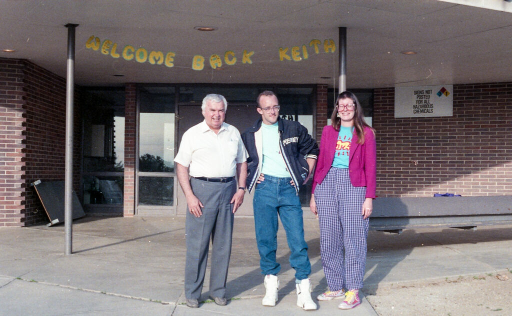 Three people stand in front of the entrance to a building under a golden banner that reads "Welcome Back Keith."