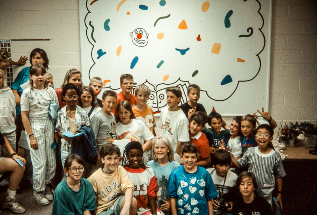 A group of about two dozen children stand smiling in front of a half-finished mural on the wall behind them. Some of the children are giving bunny ears to one another.