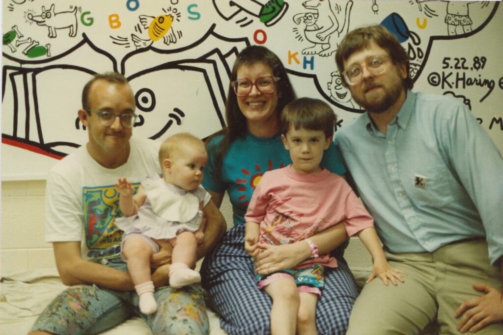 Three adults hold one small child and one baby in front of a colorful mural.