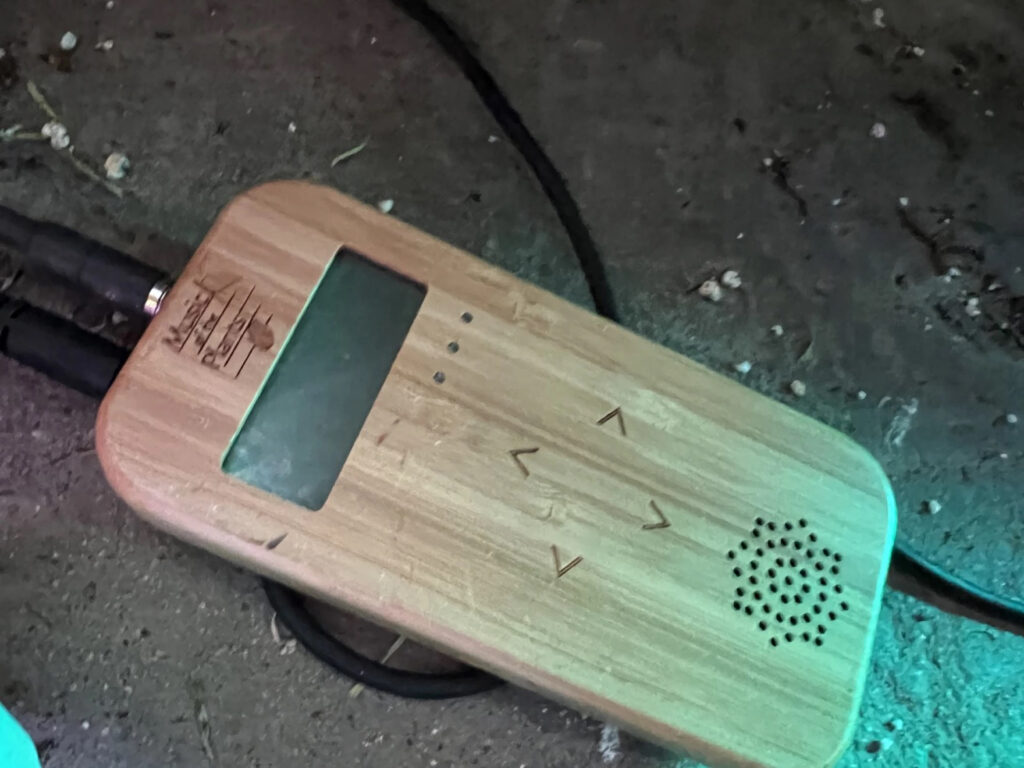 A small wooden device pictured on the ground with black cords running out of it. There is a small speaker on the bottom and a narrow rectangular digital screen at the top. In the center, four navigation arrows.