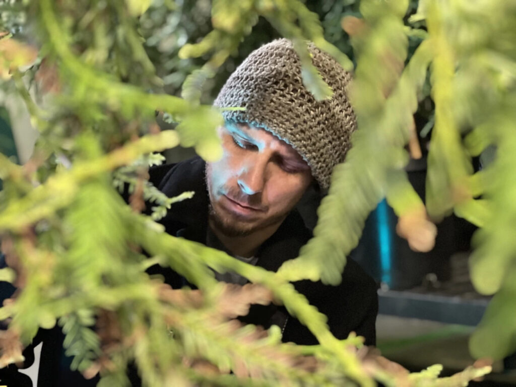 A person in a knit beanie looks down at something. Their face is framed by foliage.