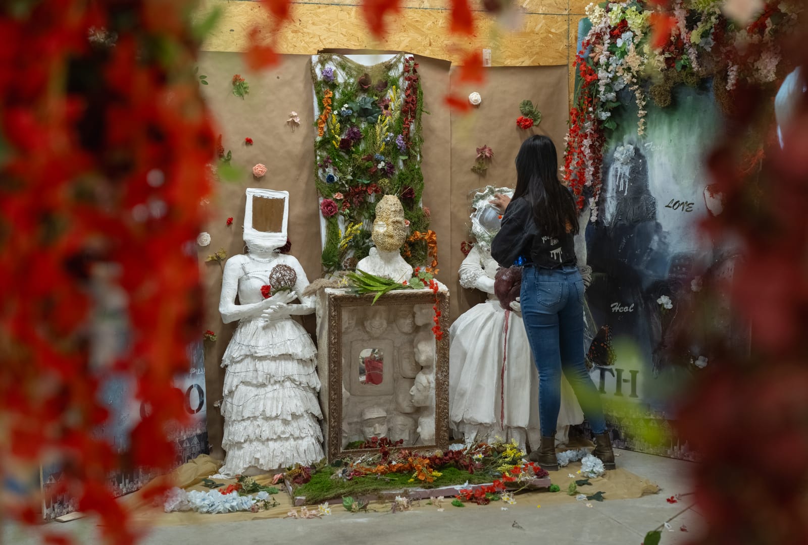 A person with long black hair has their back turned to the camera and is touching a white sculpture of a human form about a foot shorter than them. There are flowers, leaves, and another similar white sculpture strewn around the display.