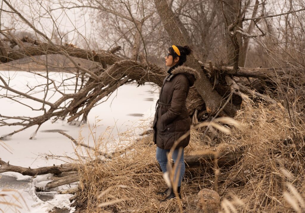 A person wearing a brown coat stands at the edge of a frozen pond. Brown branches and trees dip into the pond.