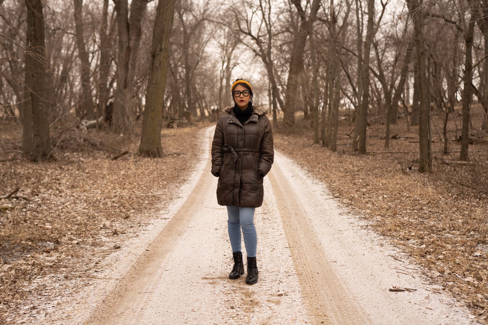 A person wearing a brown jacket stands in the center of a dirt road lightly dusted with snow. They are surrounded by a forest of leafless trees.