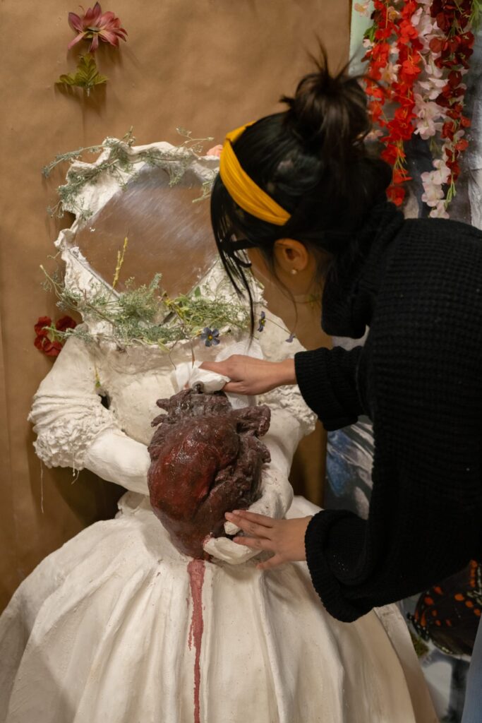 A person wearing a black turtleneck adjusts a large bleeding heart sculpture that is in the center of a white sculpture of a human wearing a dress. Instead of a face, the sculpture has a mirror its head framed with pine needles and branches.
