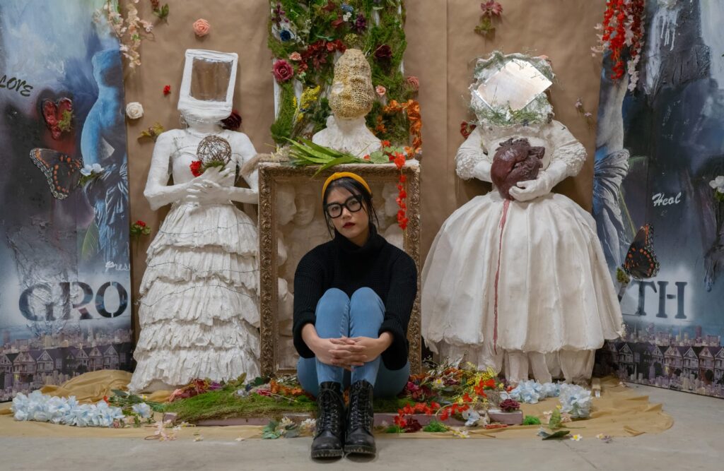 A person with long black hair pulled up wearing thick black glasses and a black turtle neck sits with their knees pulled to their chest in between two white sculptures.