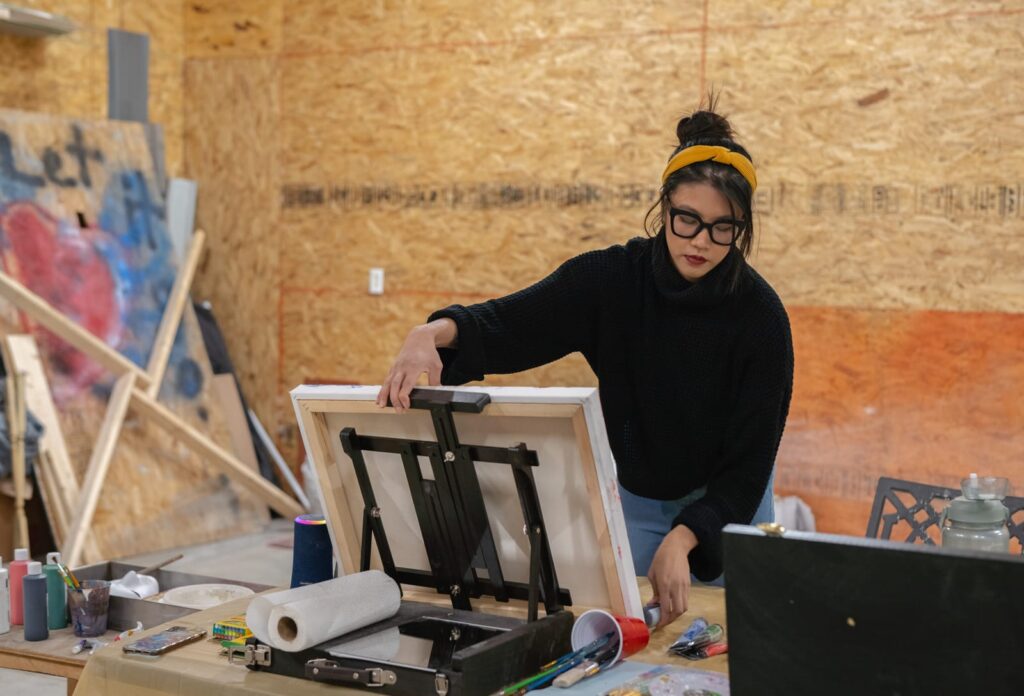 A person wearing a black turtle neck and black thick framed glasses adjusts an easel that is on the table in front of them.
