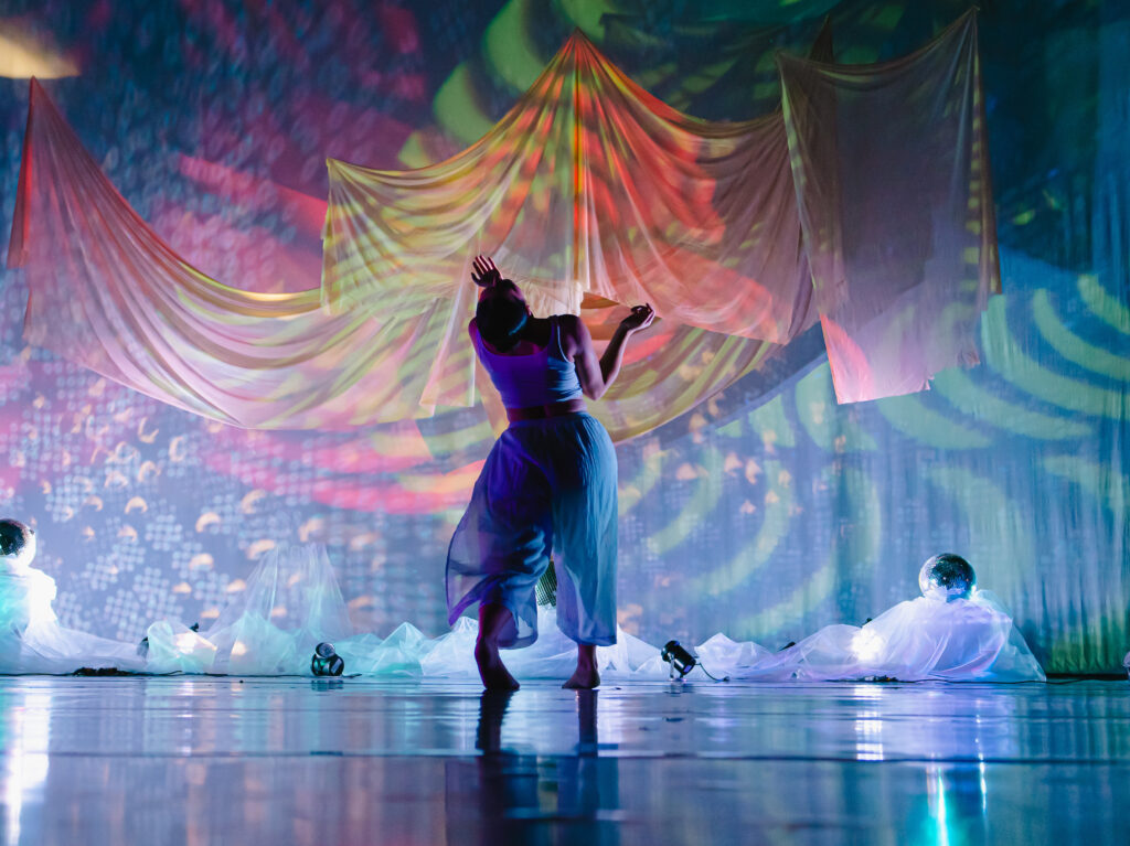 ​​A dancer wearing a cropped tank top and long flowy pants, on a stage with draped fabrics, disco balls, and colorful projections covering the walls.​