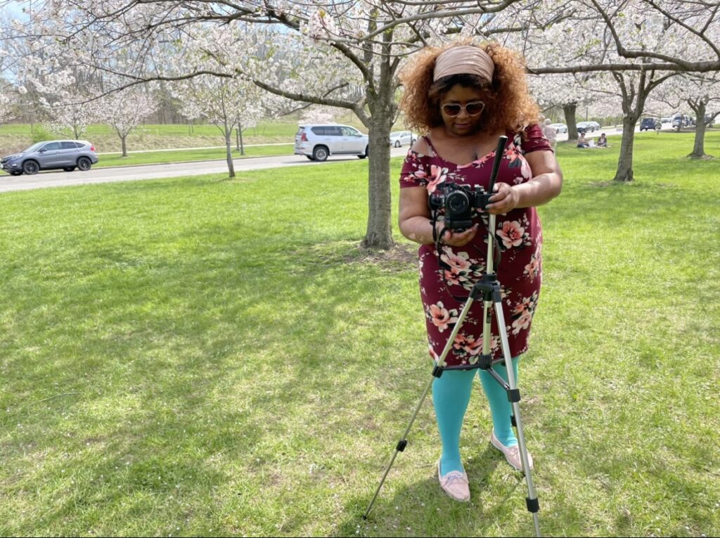 A person wearing big sunglasses and a colorful outfit adjusts a camera on top of a tripod in a grassy area surrounded by flowering trees.