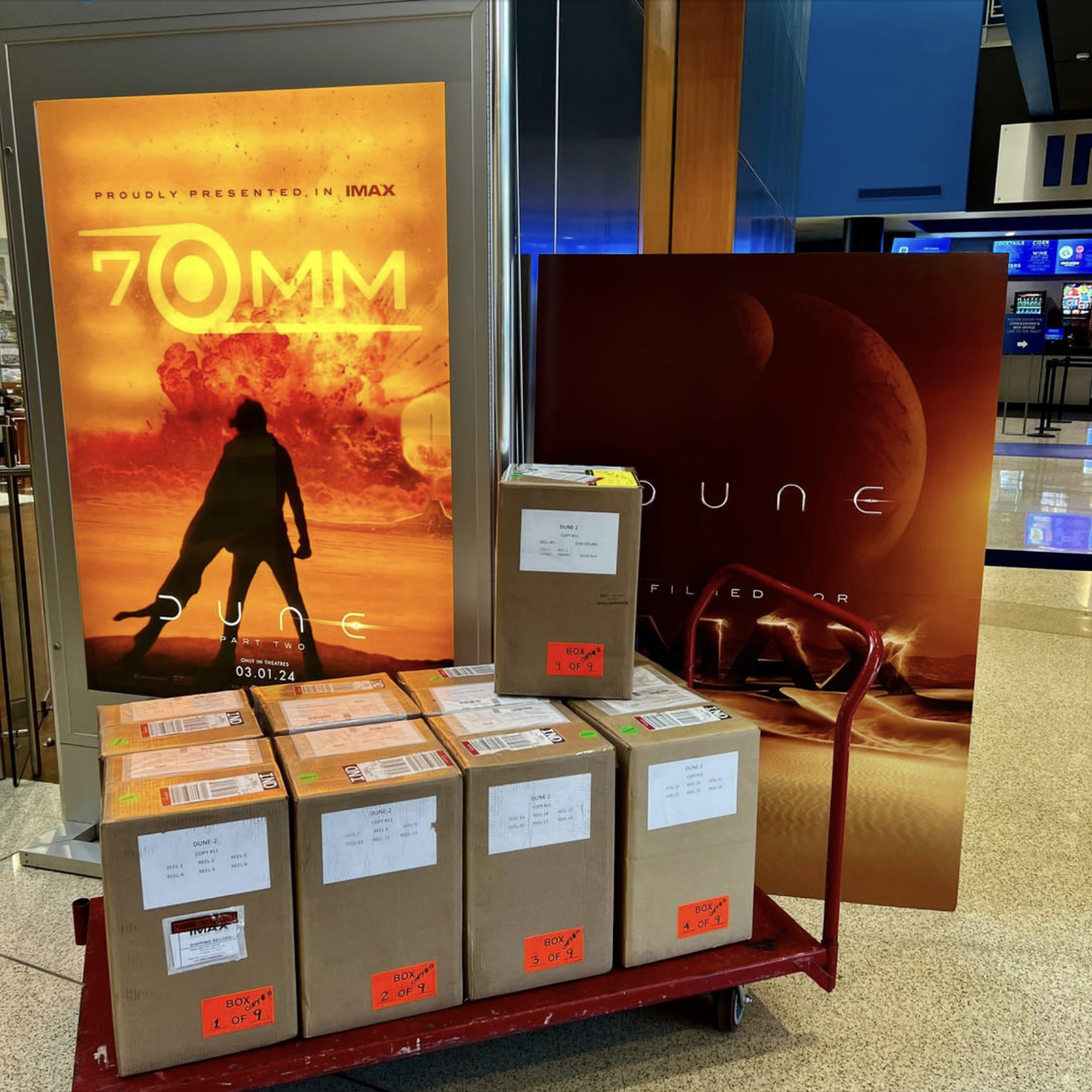 A tray cart carrying nine boxes, in front of a large display advertising Dune Part Two in 70MM IMAX.