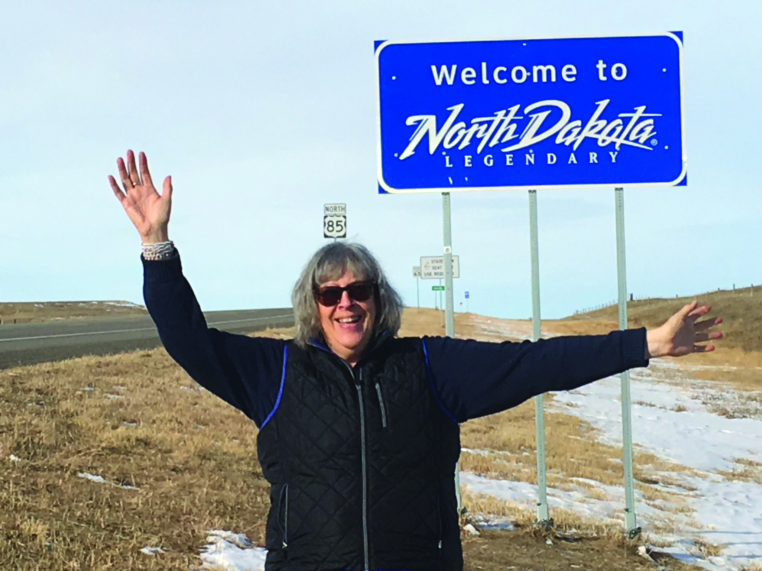 A woman waves in front of a sign that reads Welcome to North Dakota