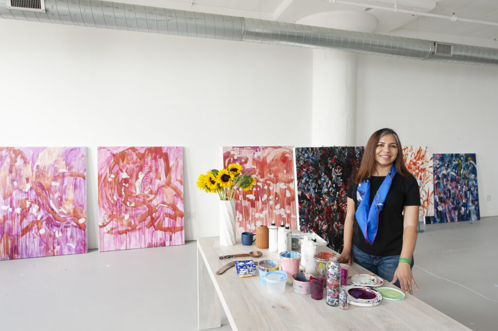 A person of medium skin tone and long brown hair stands by a long table and smiles. They are wearing a bright blue scarf around their neck, a black tshirt, and denim pants. In the background, there are multiple large paintings against a large white wall.