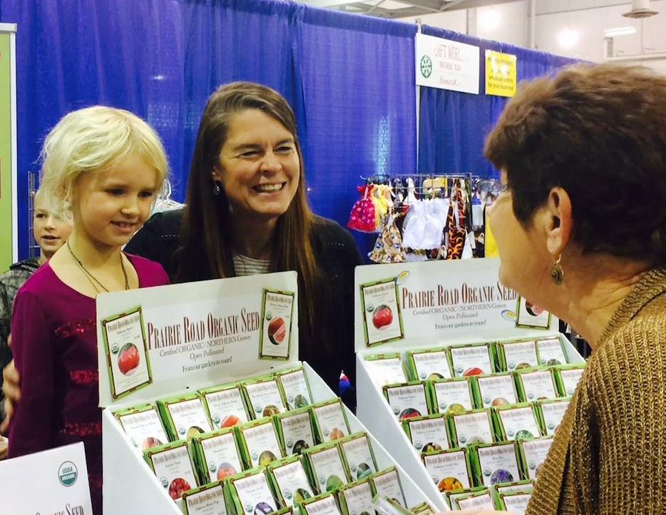 A woman of light skin tone has a young child standing by them as they talk to another person. In front of them is a display of seed packets and signage that reads, "Prairie Road Organic Seed."