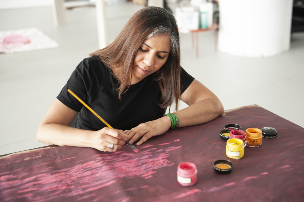 A person of medium skin tone with long brown hair adding paint to an artwork laid out on a table. They are holding a long yellow paintbrush and there are paint jars by them.