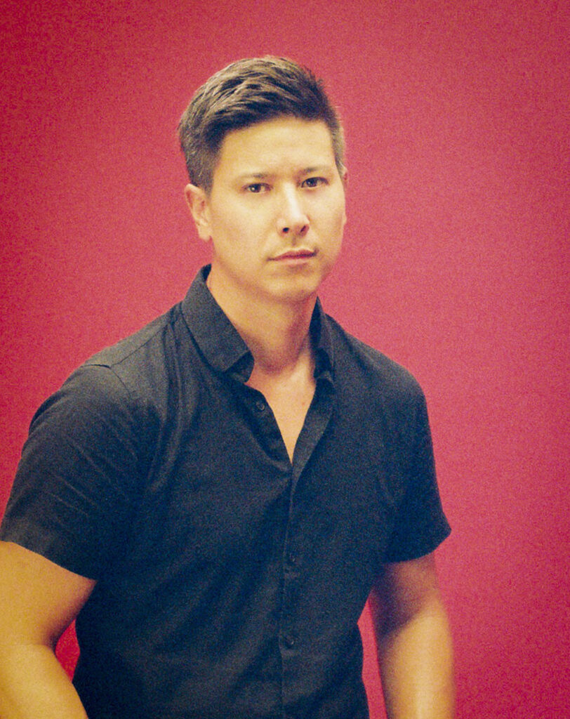 A person of medium skintone, short hair, and a black collared shirt standing in front of a red backdrop.