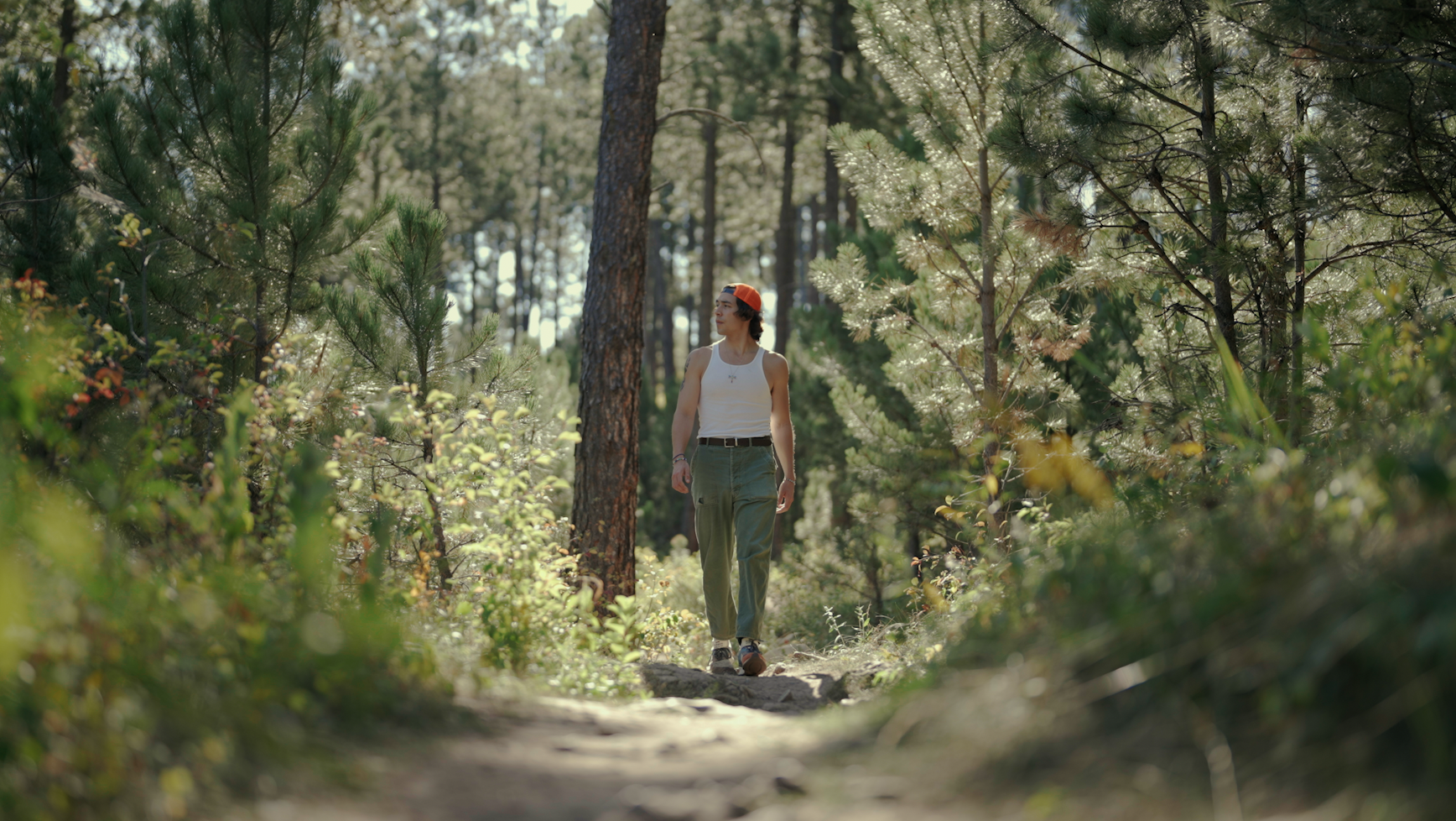 A person of medium skin tone wearing green cargo, a white tank top, and an orange bandana on their head walking in a forest area.