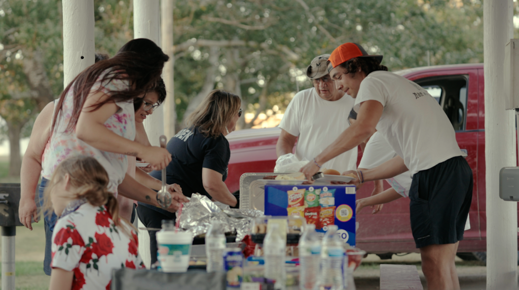 A group of people gather around a table with bottles of water and other drinks, and food items at an outdoor shelter.
