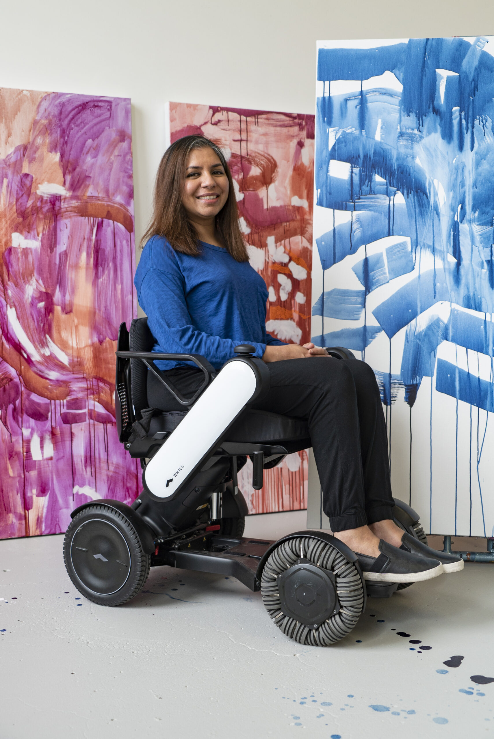 A person of medium skin tone and long brown hair using a wheelchair smiles and poses in front of large colorful paintings. They are wearing a blue long-sleeve shirt, black pants, and black slip-on shoes.