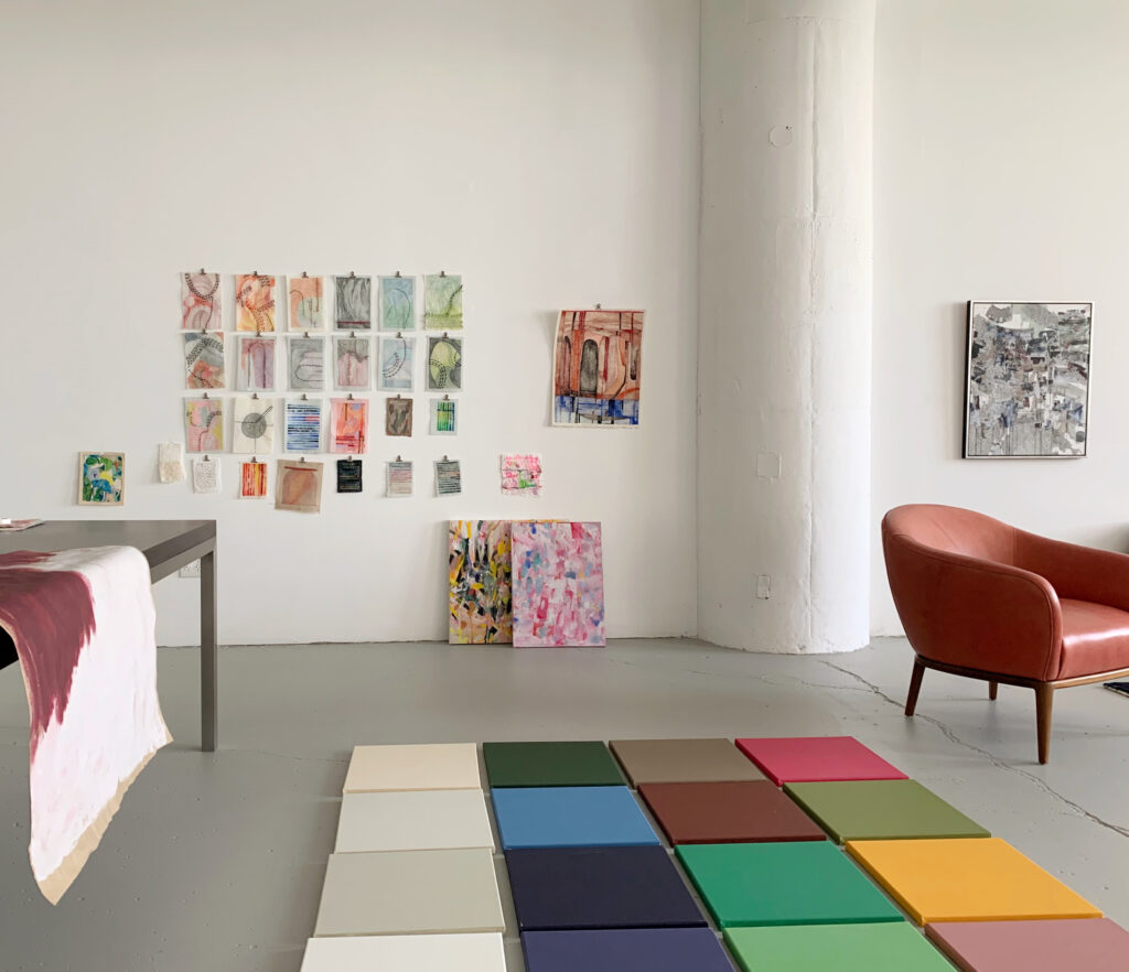 A wide view of a room with white walls. There are artworks of varying sizes on the walls and a colorful collection of square canvases on the floor.