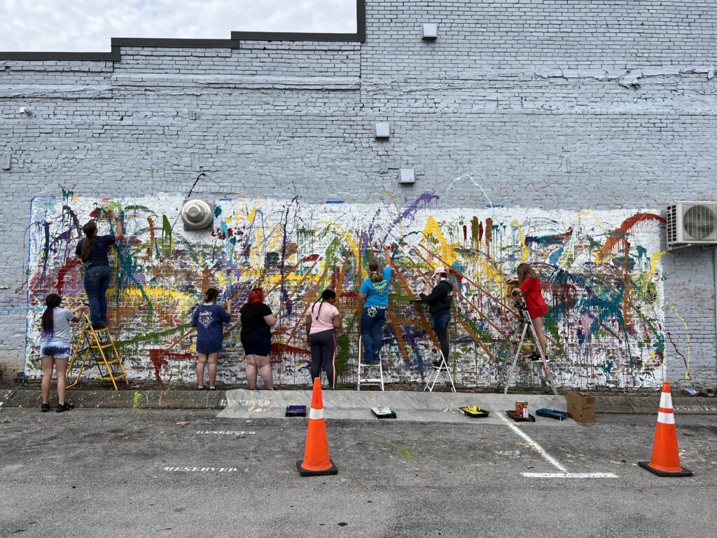 A group of students paint colorful streaks on a boring wall.