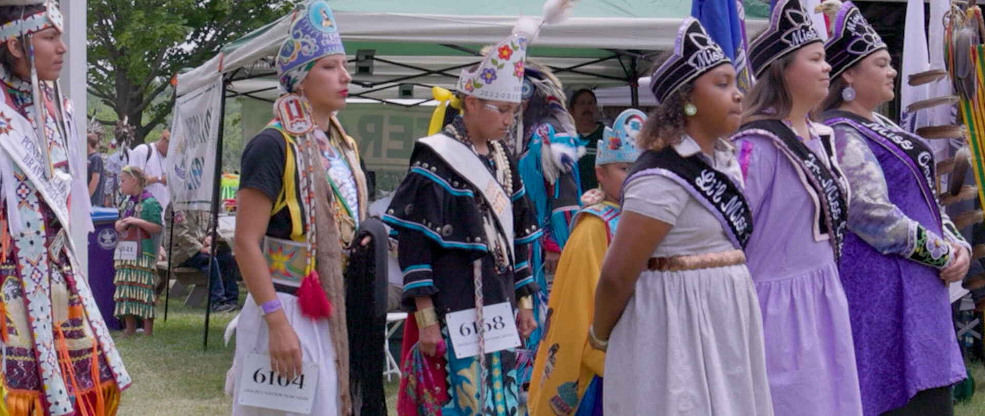 A group of young people wearing traditional Native American regalia as well as pageant style sashes. Some of them have paper with competition numbers pinned to their skirts.