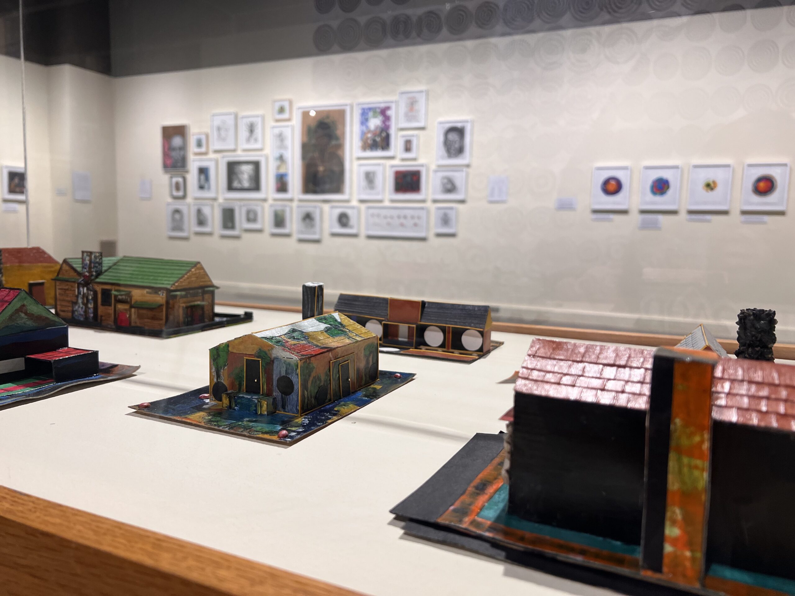 An art exhibition with a display of miniature house sculptures. The wall in the background has varying sizes of framed drawings and paintings hung on it.