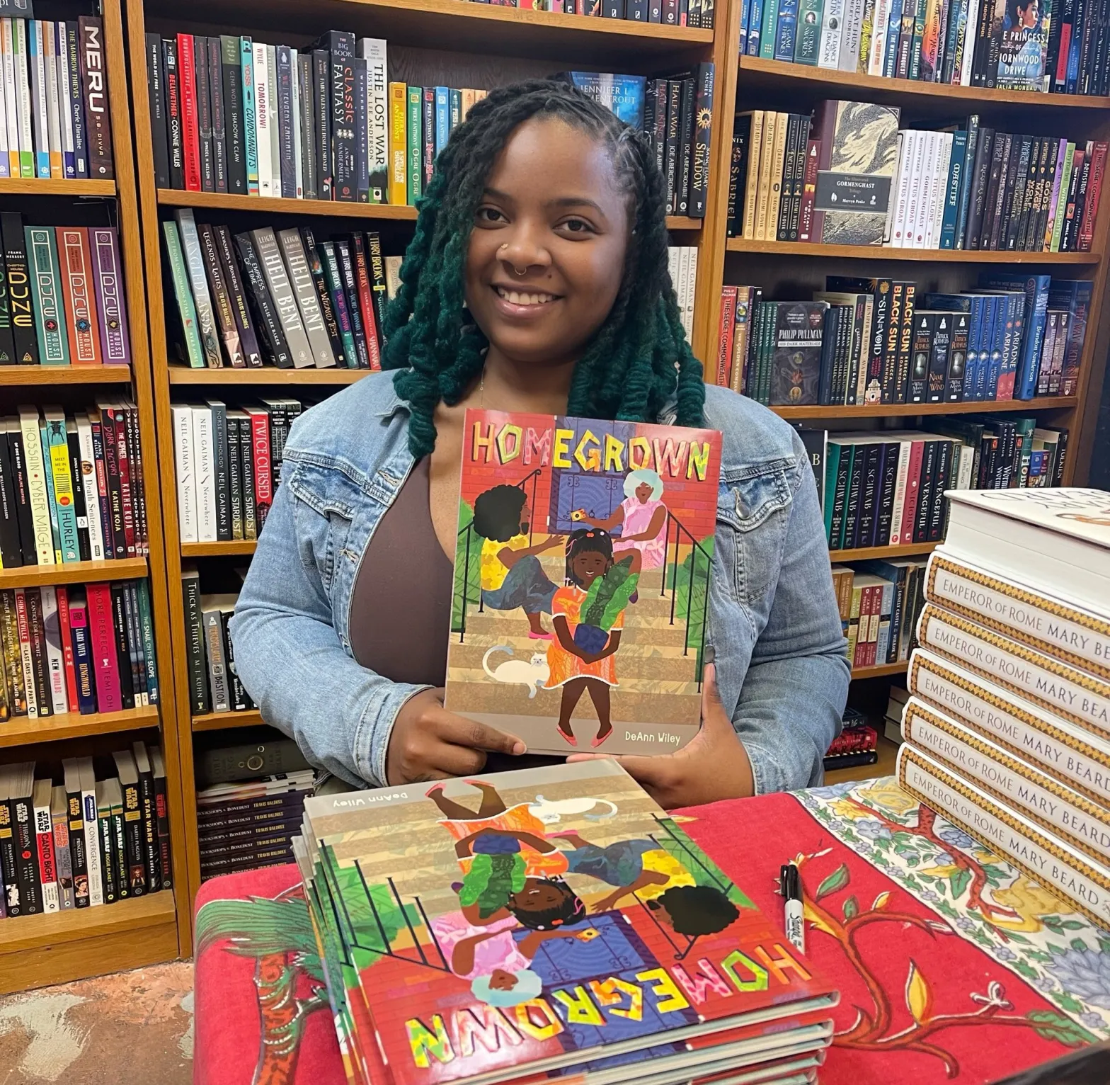 A person with teal braids and a light denim jacket holding up a book standing in front of a shelf of more books.