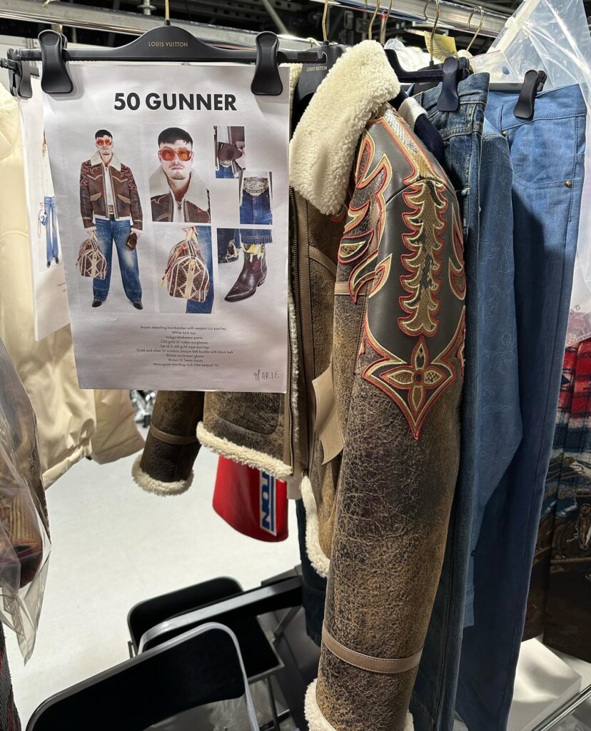 A clothing rack with a leather jacket and a few pairs of denim jeans on hangers. There is a sheet of paper that has "50 Gunner" written on it with photos of a person modeling the items on the rack.