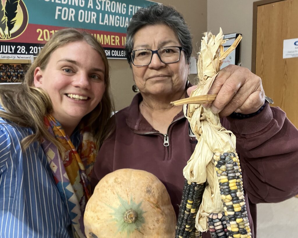 Two people stand together looking at the camera. One holds a large beige squash and the other holds the cream colored leaves of three ears of black and yellow corn between their fingers.