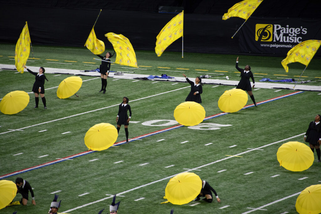 Performers on a football field. They are wearing black dresses and have bright yellow umbrellas and flags as props.