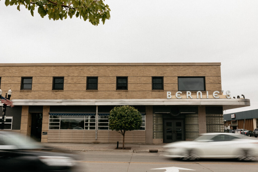 A two story blonde brick building including metal letters its awning above the door spelling "Bernie's." Two cars pass in front of the building. The sky is steely grey but trees in the foreground have green leaves.