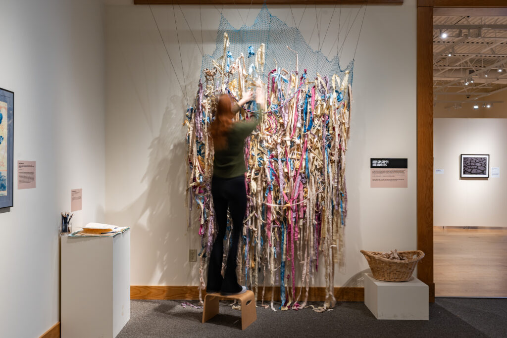 A person standing on a stepstool with their hands reaching up to the top of a fiber art piece consisting of blue woven netting and fabric strips of various colors, next to gallery signage reading, "Mississippi Memories."