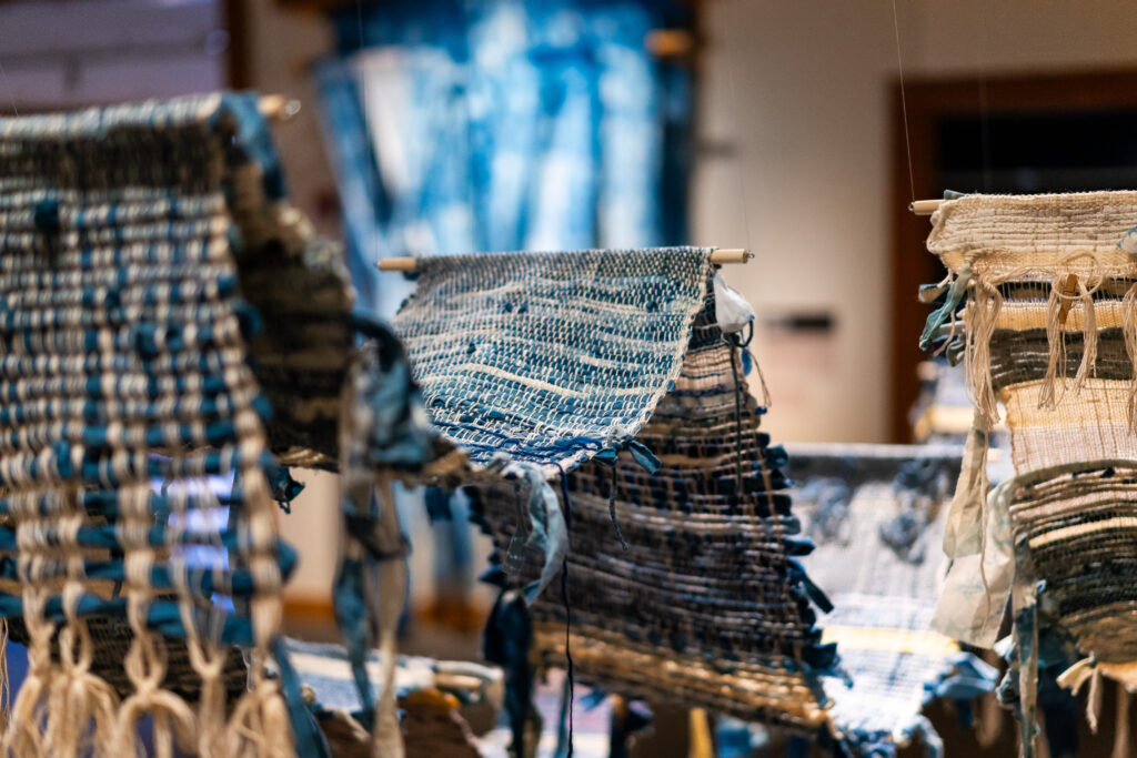 A long blue woven tapestry held up by wooden rods, hung up in a gallery space.