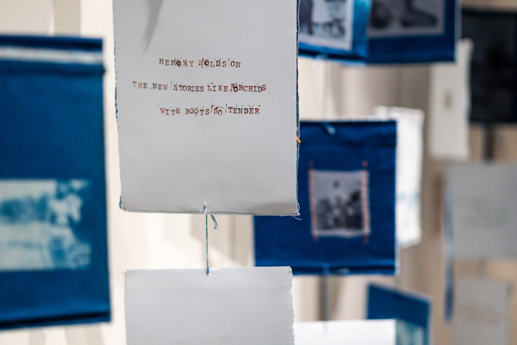 A fiber and print artwork, with text reading, "memory holds on, the new stories like orchids, with roots so tender" surrounded by several fabric pieces with cyanotype prints.