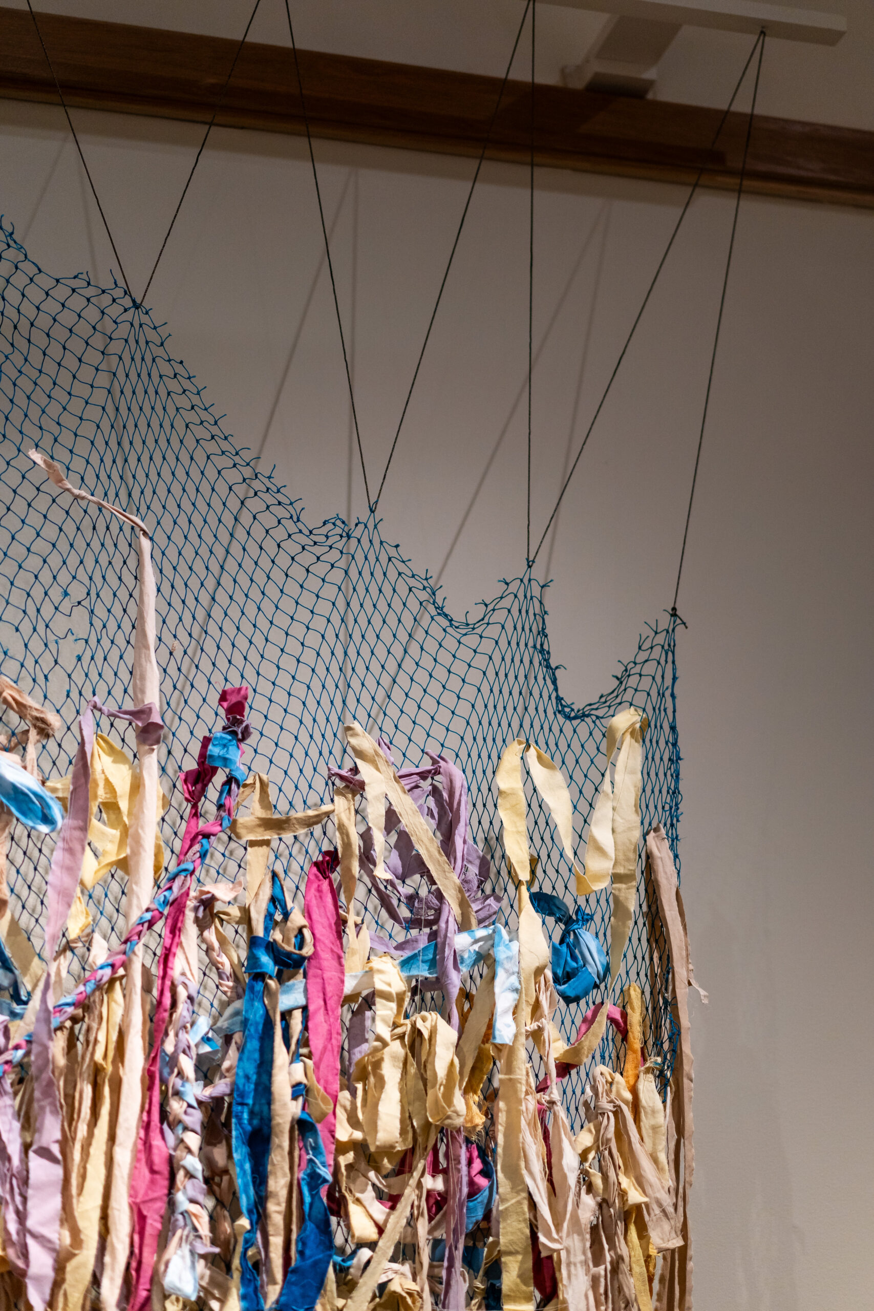 A fiber art piece hung from the ceiling, consisting of fabric strips of various colors tied into a section of blue woven netting.